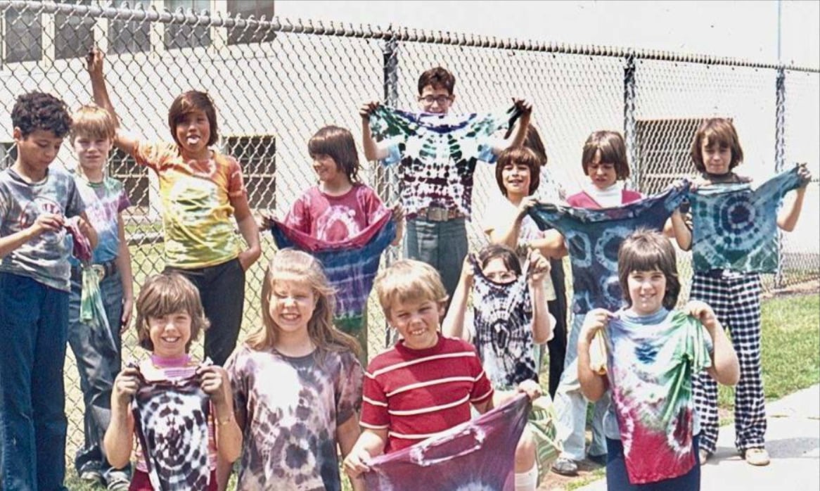 A group of children stand outside in front of a chain-link fence, smiling and holding up colorful tie-dyed shirts on a sunny day.