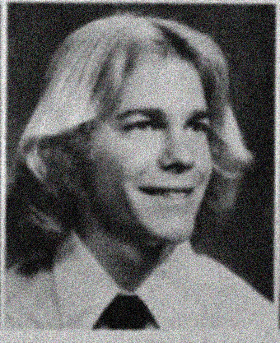 Black and white yearbook-style portrait of a young person with shoulder-length, light-colored hair, wearing a collared shirt and dark tie, smiling and looking slightly to the side.