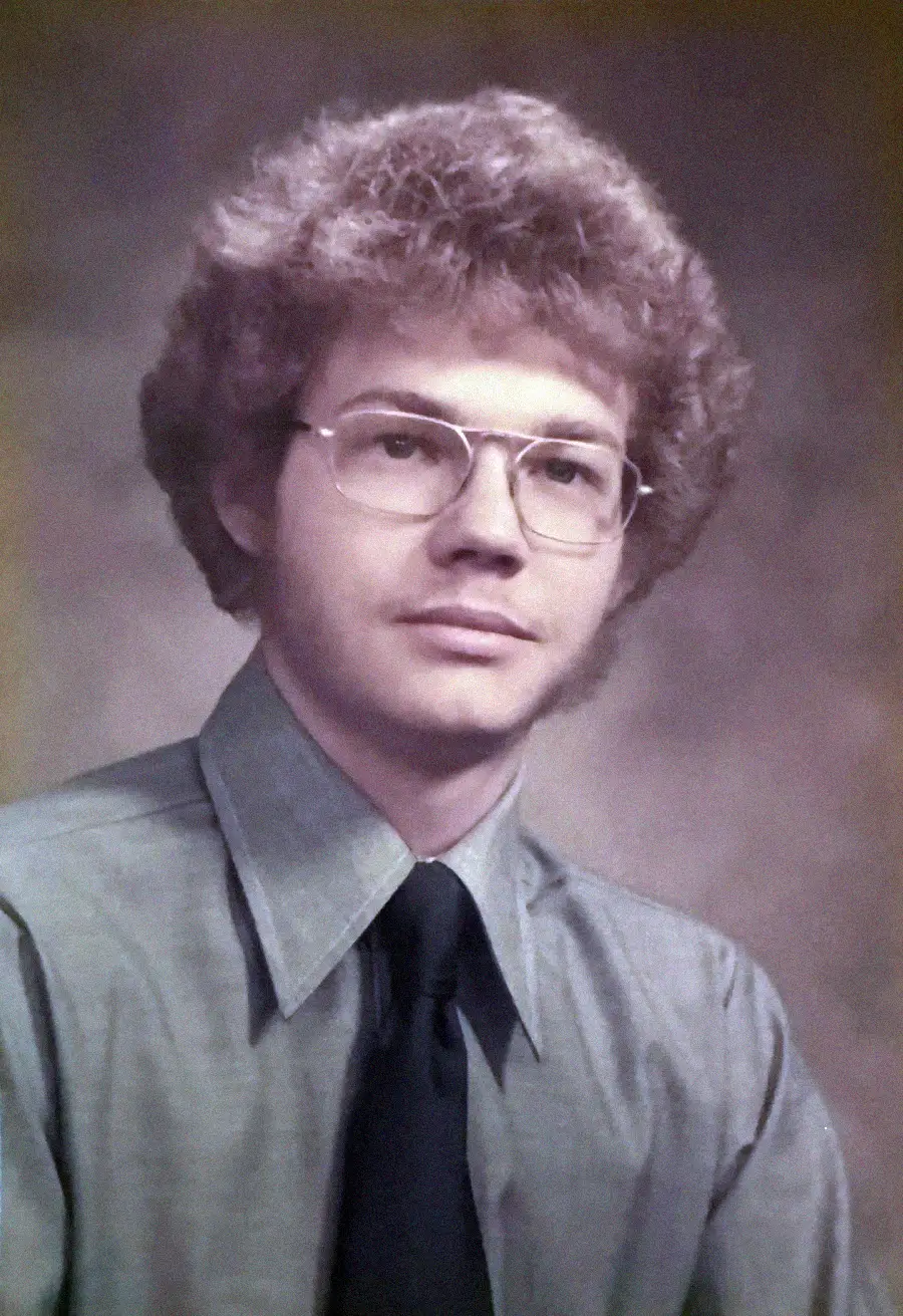 A young man with curly hair, glasses, and sideburns, wearing a light gray shirt with a wide collar and a dark tie, poses for a formal portrait against a neutral background.