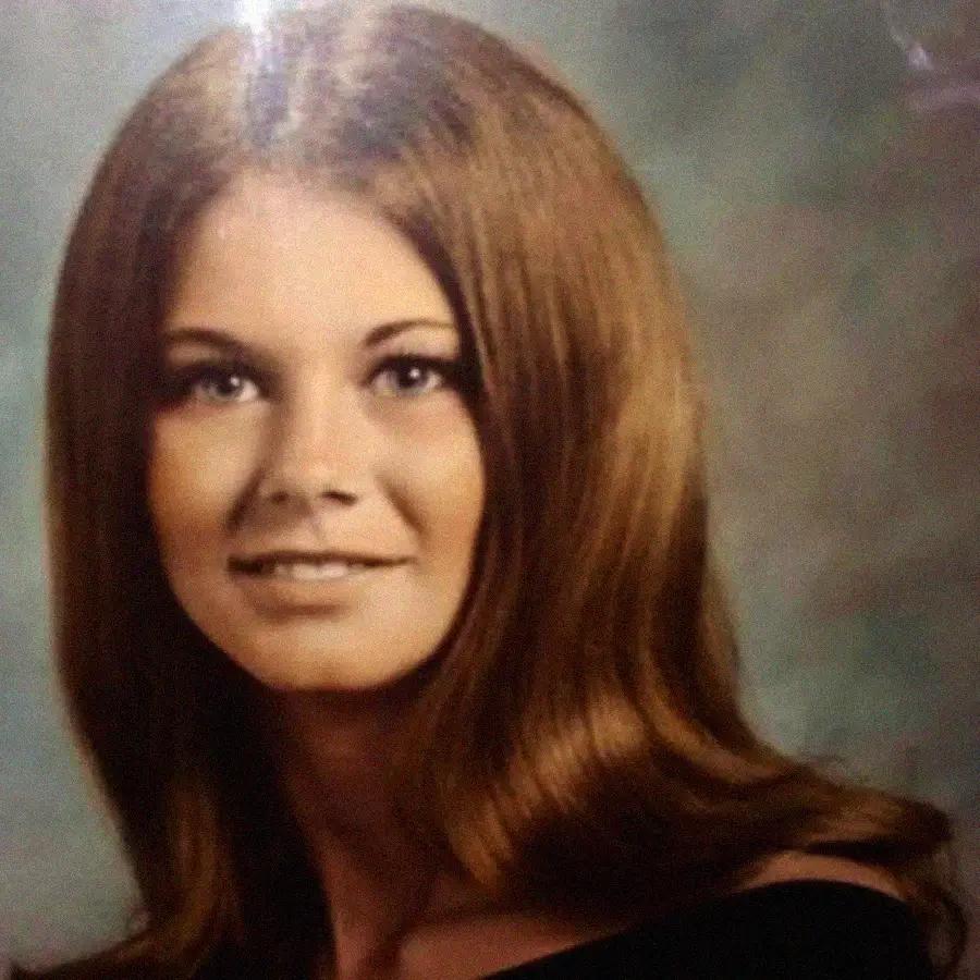 A young woman with long, straight brown hair and a slight smile poses for a formal portrait in front of a blurred background, wearing an off-the-shoulder black top.
