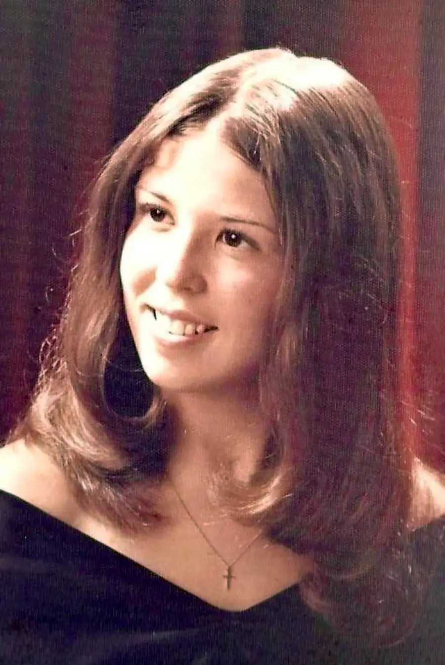 A young woman with long brown hair, wearing an off-the-shoulder black top and a cross necklace, smiles softly while looking slightly to the side against a dark reddish background.