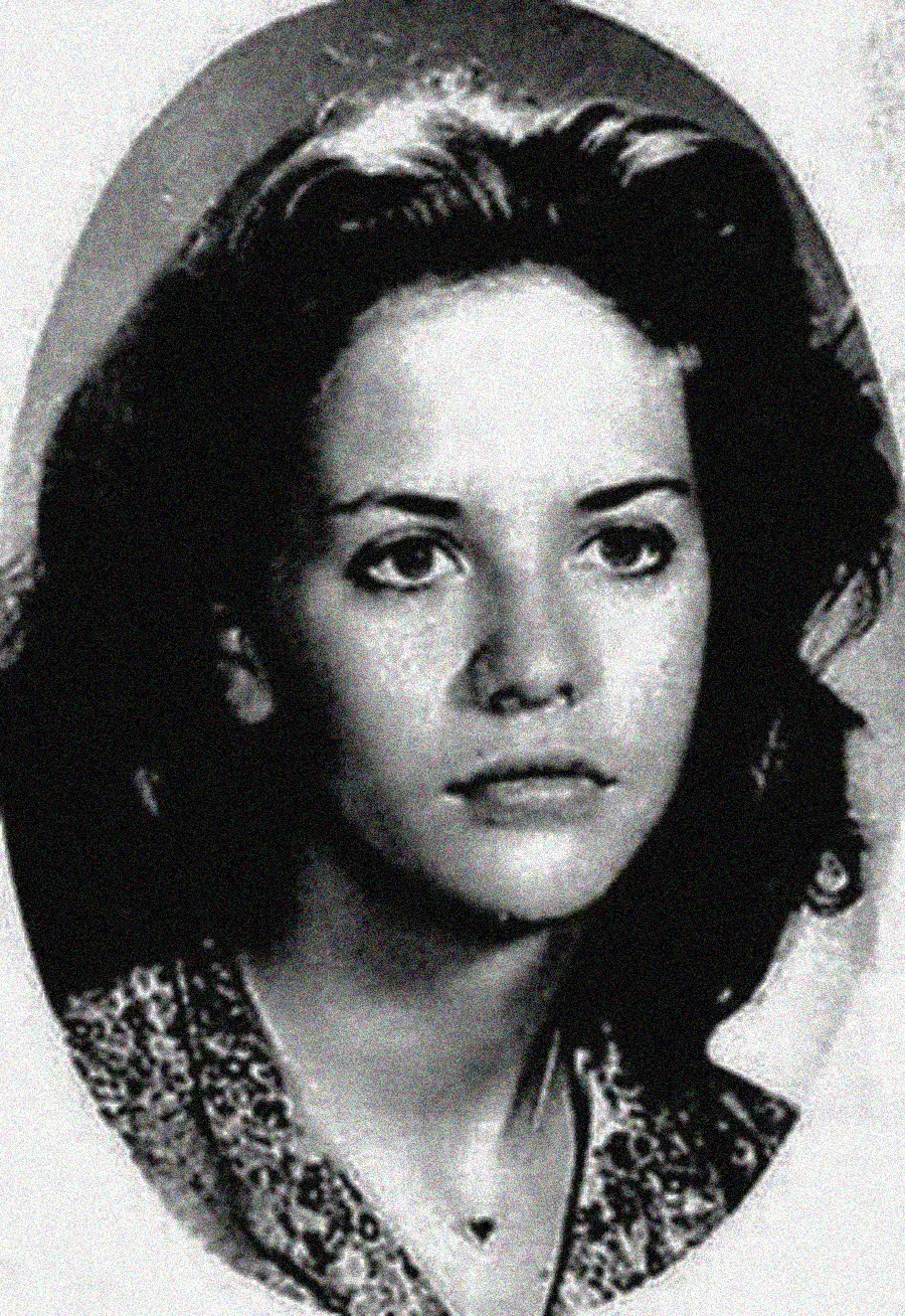 A black and white portrait of a young woman with wavy hair, looking slightly to the side with a neutral expression, wearing a patterned top and a small necklace. The photo has a soft, vintage appearance.