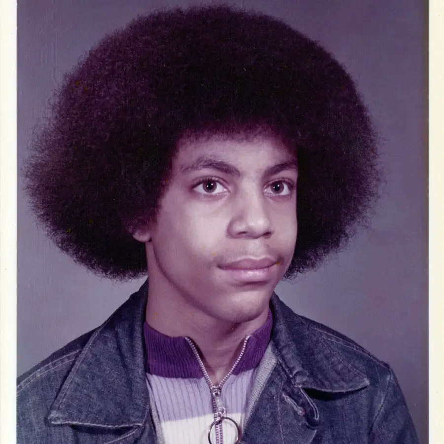 A young person with a large afro hairstyle, wearing a denim jacket over a zip-up shirt with a ring zipper pull, poses for a studio portrait against a plain, neutral background.