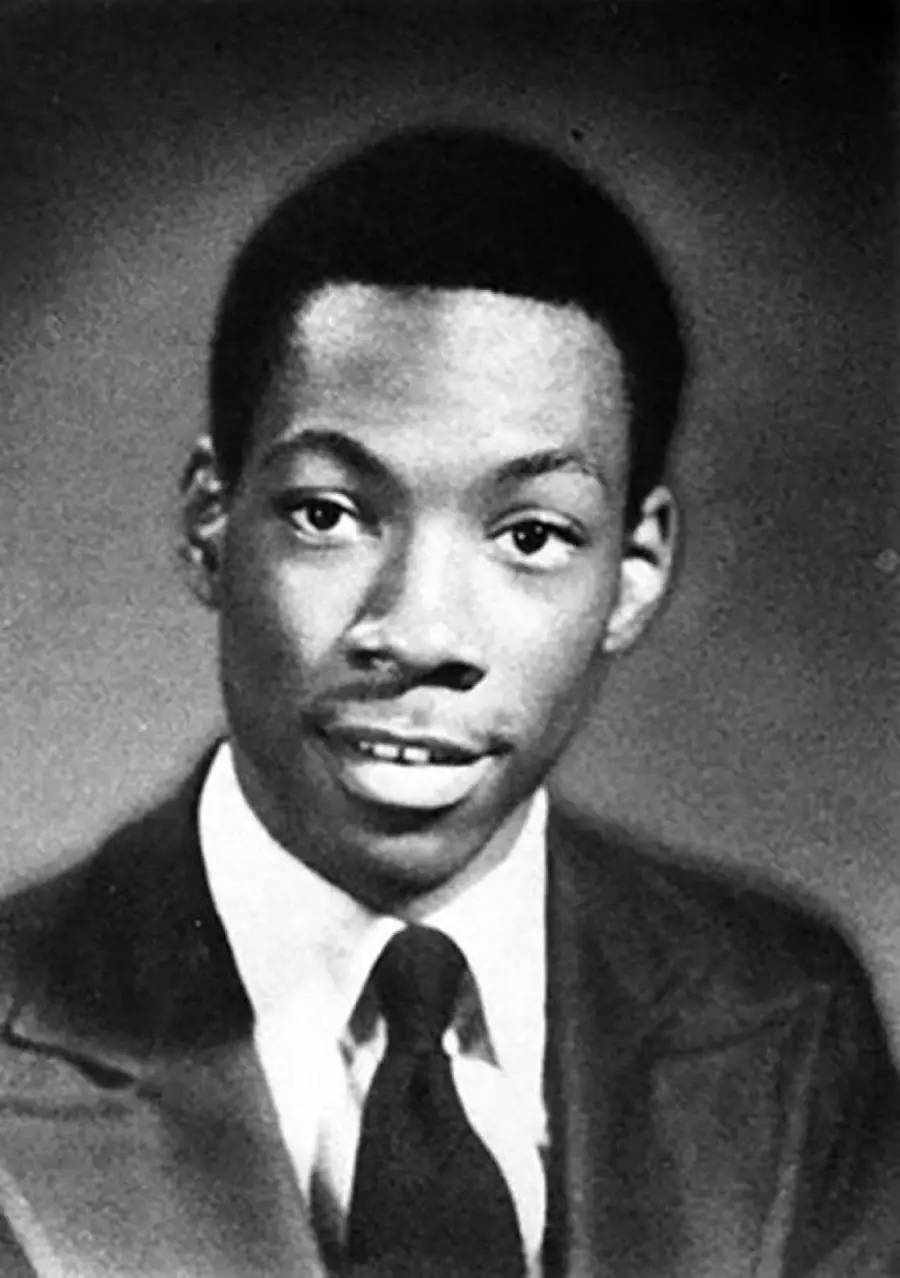 A young man wearing a suit and tie poses for a formal black-and-white portrait against a dark background. He faces forward and smiles slightly.