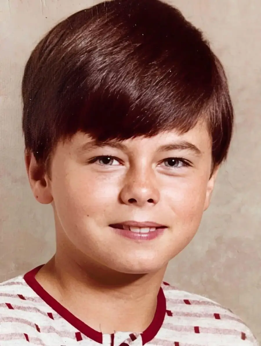 A young boy with straight brown hair and fair skin smiles softly, wearing a white shirt with red stripes, in front of a plain beige background.