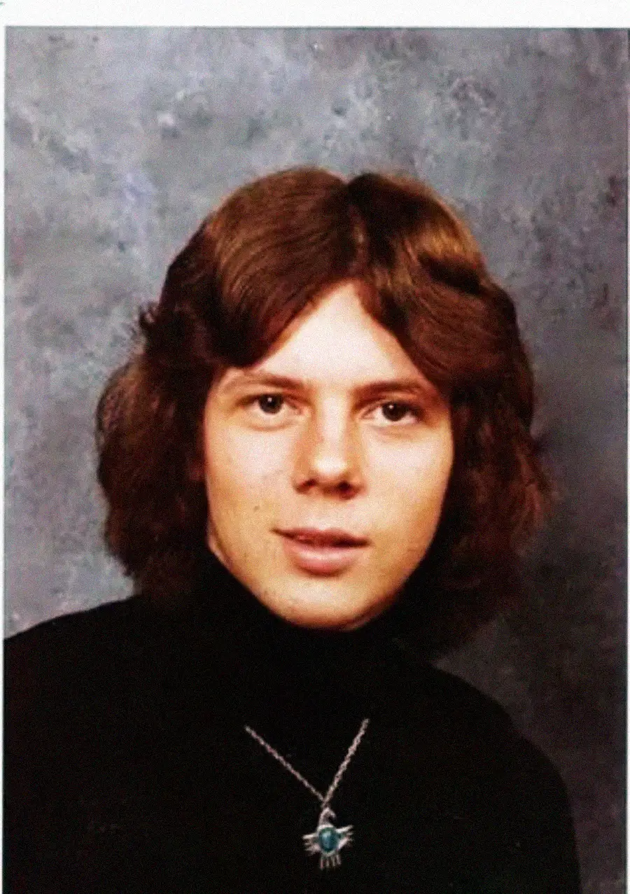A young person with medium-length brown hair wearing a black turtleneck and a necklace with a turquoise pendant poses in front of a mottled gray background.