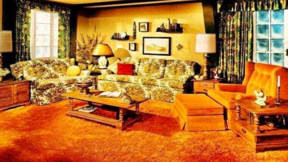 A retro living room with floral-patterned sofas, an orange armchair, orange shag carpet, wood furniture, and floral curtains. The room is decorated with lamps, plants, and framed pictures on the yellow walls.