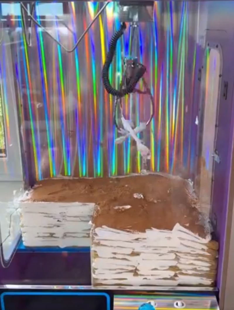 A claw machine with a holographic background contains stacks of what appear to be paper-wrapped blocks topped with brown powder, resembling tiramisu cake. The claw is positioned above the blocks, ready to grab one.
