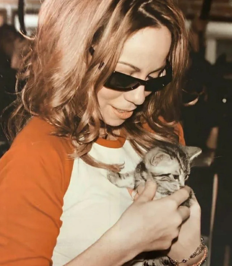 A woman with wavy hair and sunglasses smiles while gently holding a small gray tabby kitten in her hands.