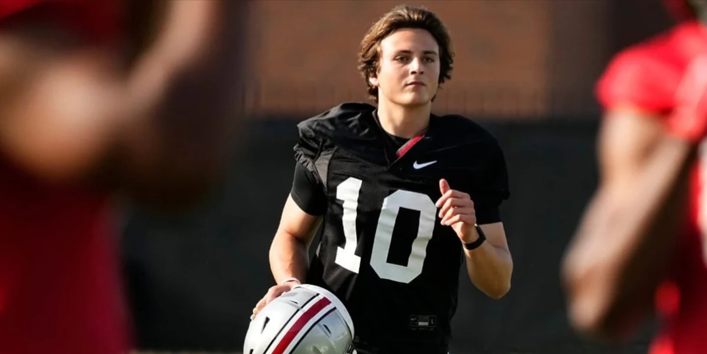 A young football player wearing a black jersey with the number 10 holds a helmet and looks ahead while jogging on a field, with other players in red jerseys blurred in the foreground.