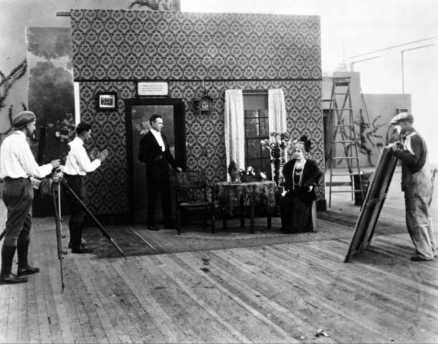 A black-and-white photo shows a film crew shooting a scene on an indoor set decorated like a living room. Two actors perform while a cameraman films, another person holds a reflector, and stage props fill the scene.