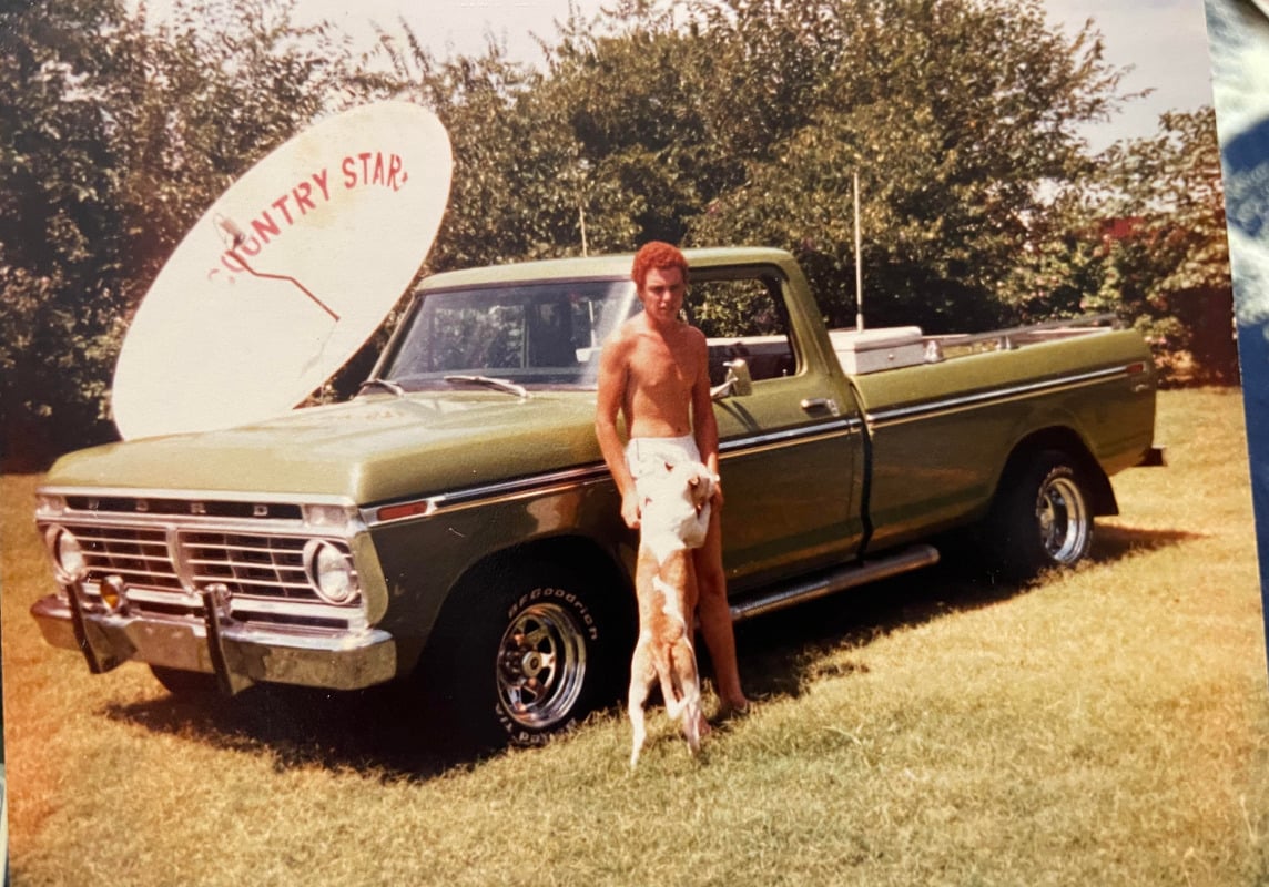 A shirtless man stands beside a green vintage pickup truck with a large "Country Star" satellite dish in the bed. A dog stands on its hind legs, leaning against the man. Trees and grass fill the background.
