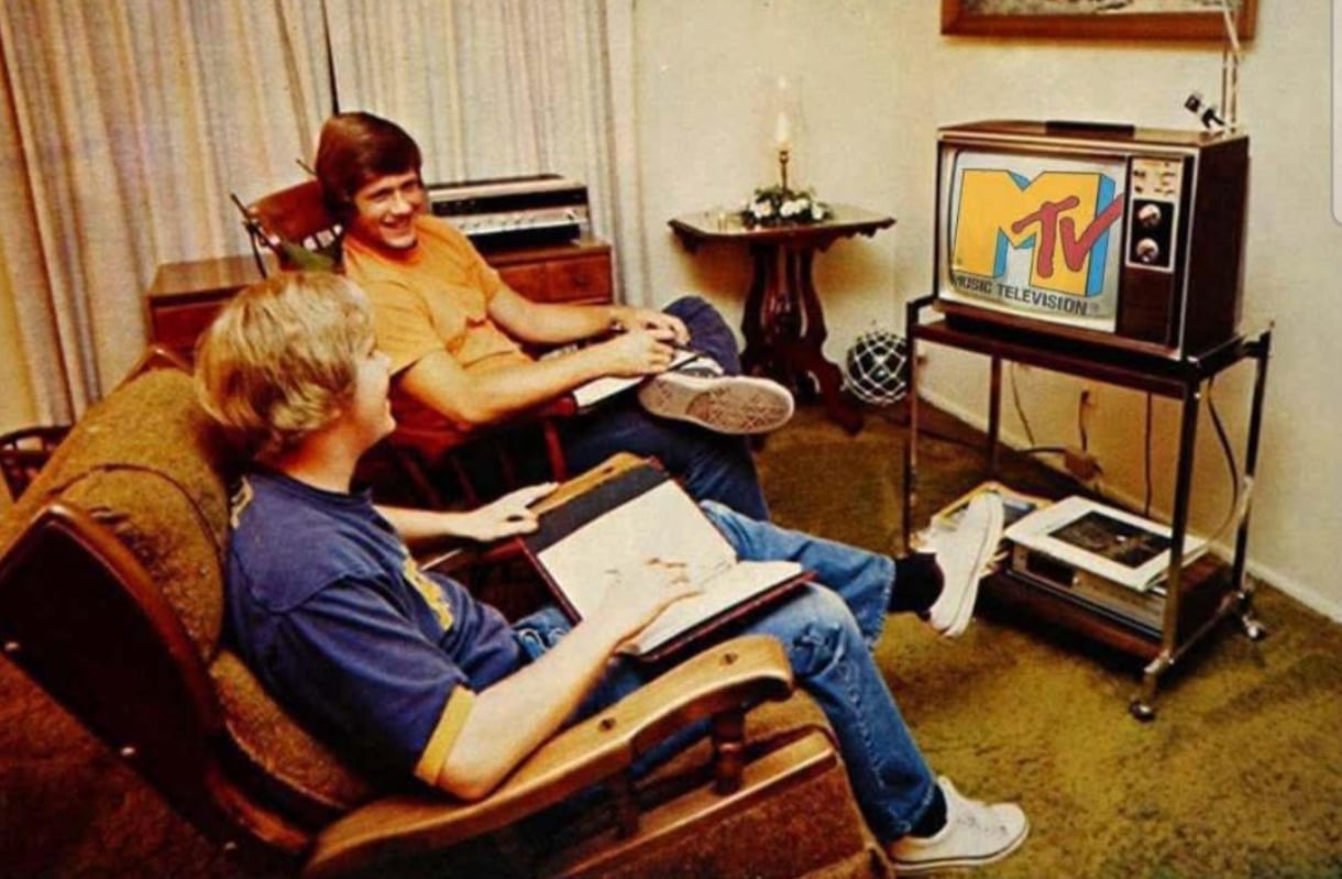 Two young men sit in a retro living room, smiling and watching MTV on an old television set. One holds a notebook, and the room features vintage decor, including a record player and shag carpeting.