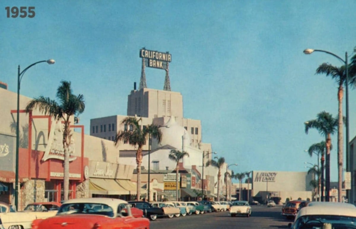 A busy street scene from 1955 shows vintage cars parked along palm-lined sidewalks with shops, restaurants, and a tall California Bank building in the background under a clear blue sky.