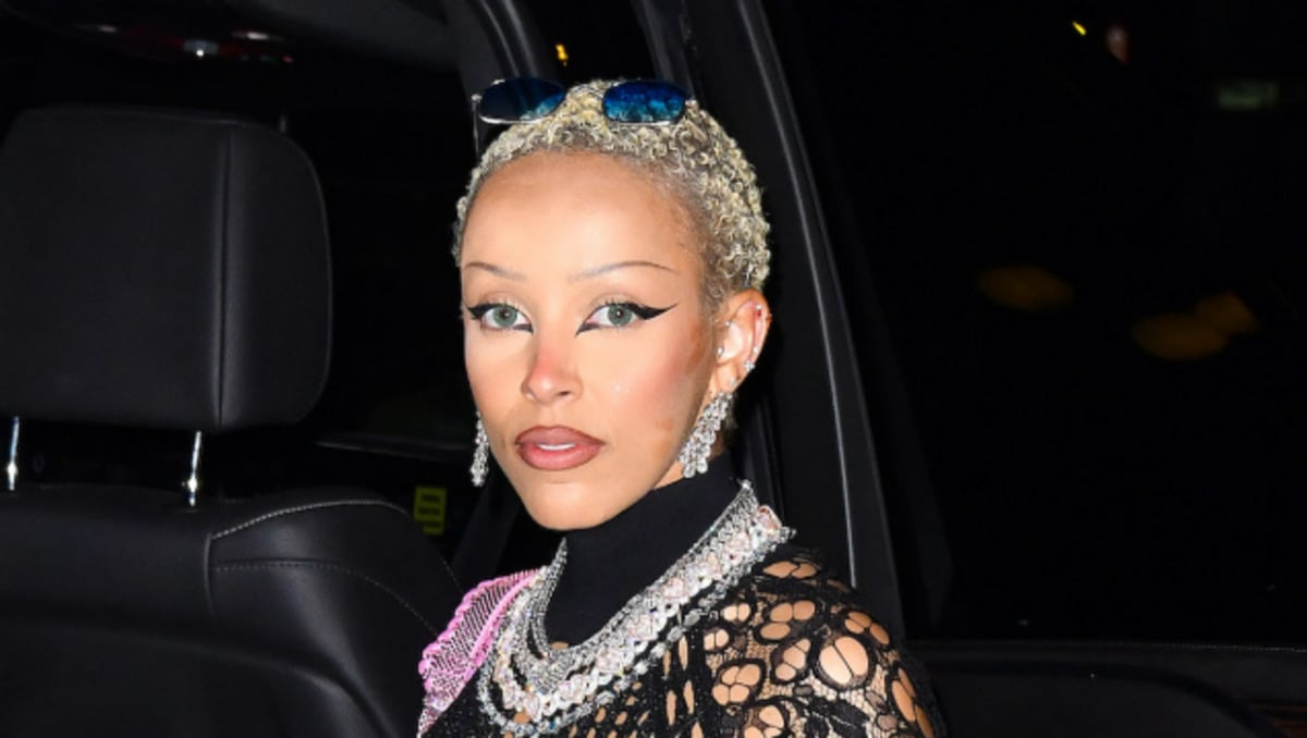 A person with short, platinum blonde hair, dramatic winged eyeliner, and statement jewelry sits in a car seat wearing a black outfit with a netted overlay and a pink accent.