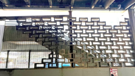 Concrete breeze blocks form a patterned wall with rectangular openings, partially revealing a staircase behind. Sunlight filters through the blocks, casting shadows. A &ldquo;No Parking Fire Lane&rdquo; sign is visible at the bottom right.
