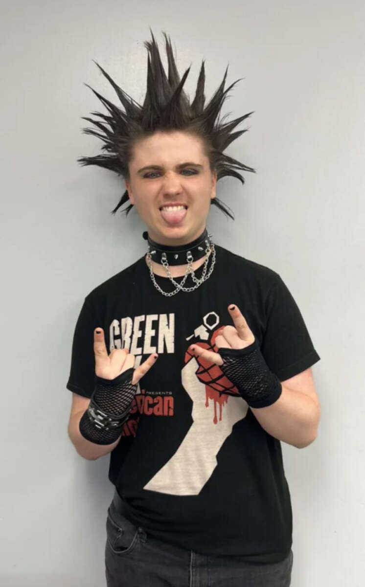A person with spiked hair, dark eye makeup, and a studded choker makes a rock hand gesture. They wear a black graphic T-shirt, black fishnet gloves, and multiple chains against a plain light background.