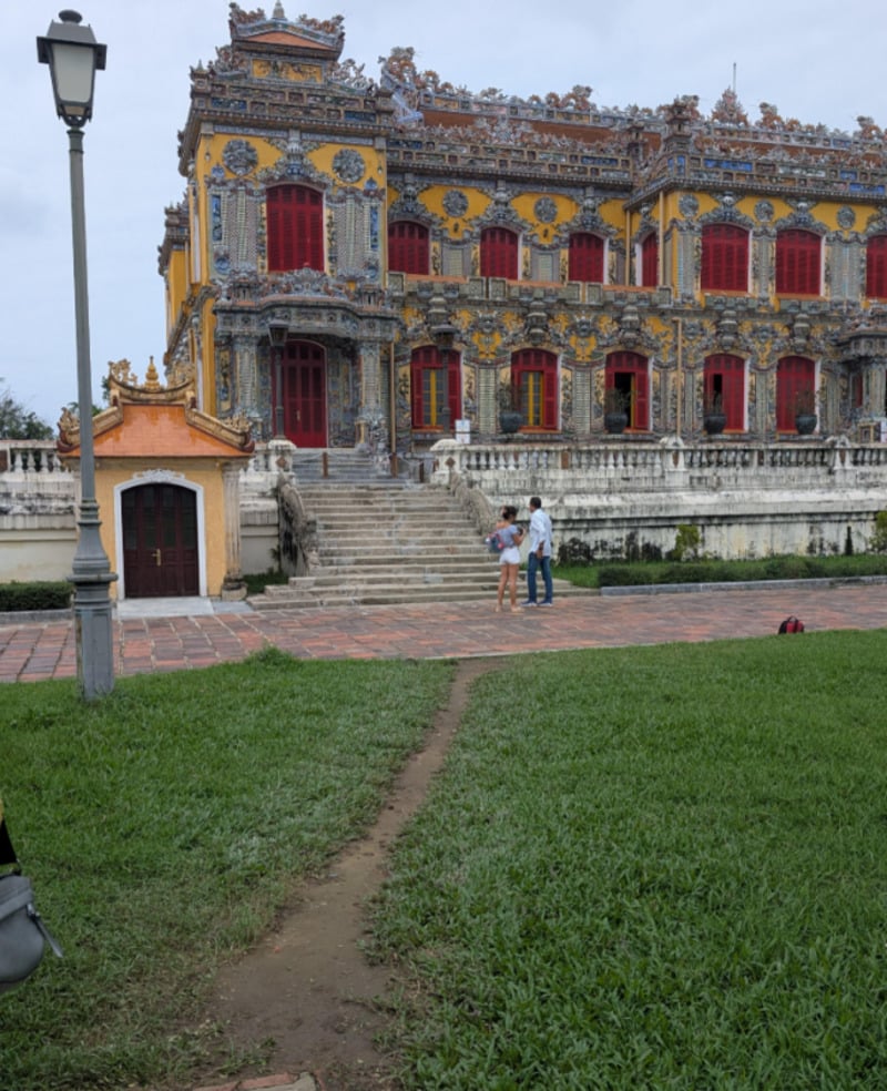 A vibrant, ornate building with yellow and blue decorations stands behind wide stone steps; two people stand at the base, and a grassy lawn with a narrow dirt path is in the foreground.