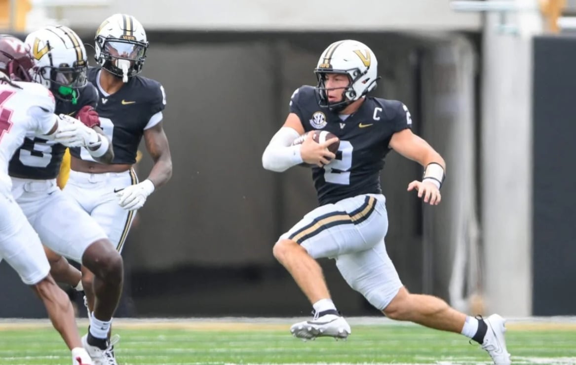 A Vanderbilt football player in a black uniform and helmet runs with the ball, evading defenders in white uniforms during a game on a football field.