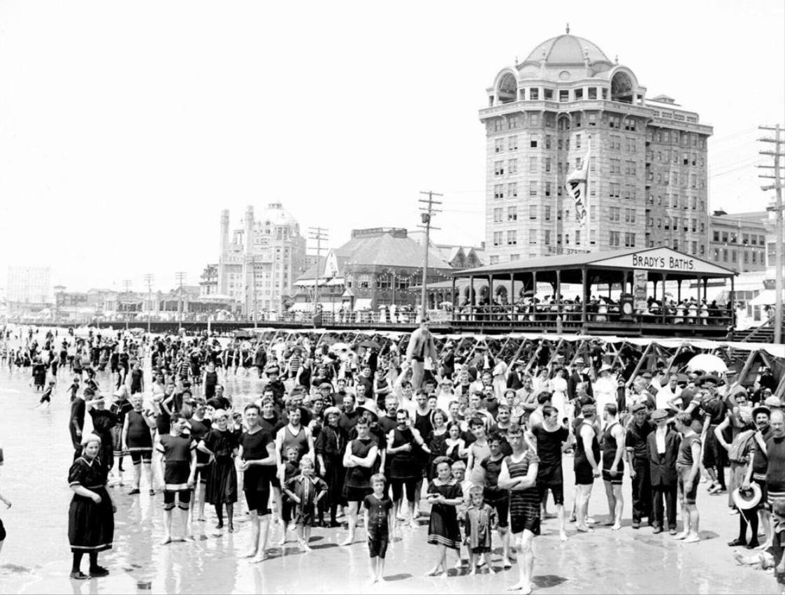 A large crowd of people in vintage swimsuits stands on a busy beach in front of a boardwalk and a grand multi-story hotel, with flags and historic buildings in the background. The scene appears to be from the early 20th century.