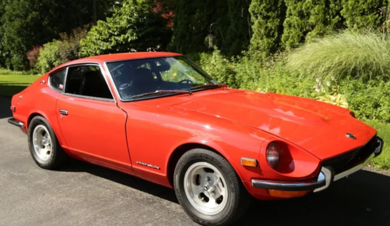 A classic red sports car, likely a Datsun 240Z, is parked on a driveway with lush green trees and bushes in the background.