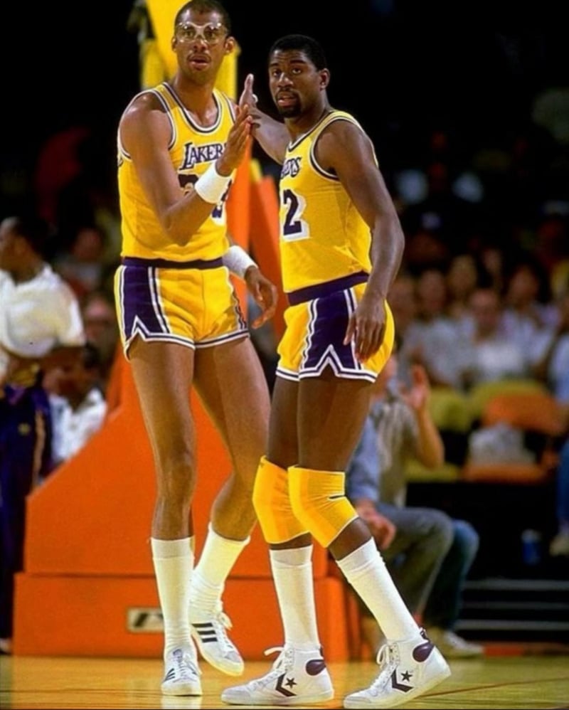 Two basketball players in yellow and purple Los Angeles Lakers uniforms stand on a court during a game, one signaling with his hand while the other looks toward the camera. Spectators are visible in the background.