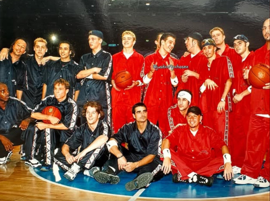 A group of young men pose on a basketball court, wearing matching black and red athletic outfits. Several are holding basketballs, and most are smiling or laughing, creating a casual, friendly atmosphere.