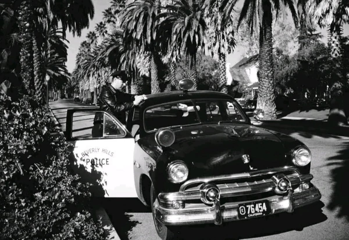 A vintage Beverly Hills police car is parked on a palm tree-lined street as an officer, in uniform and cap, speaks to someone through the driver's side window in a classic black-and-white scene.