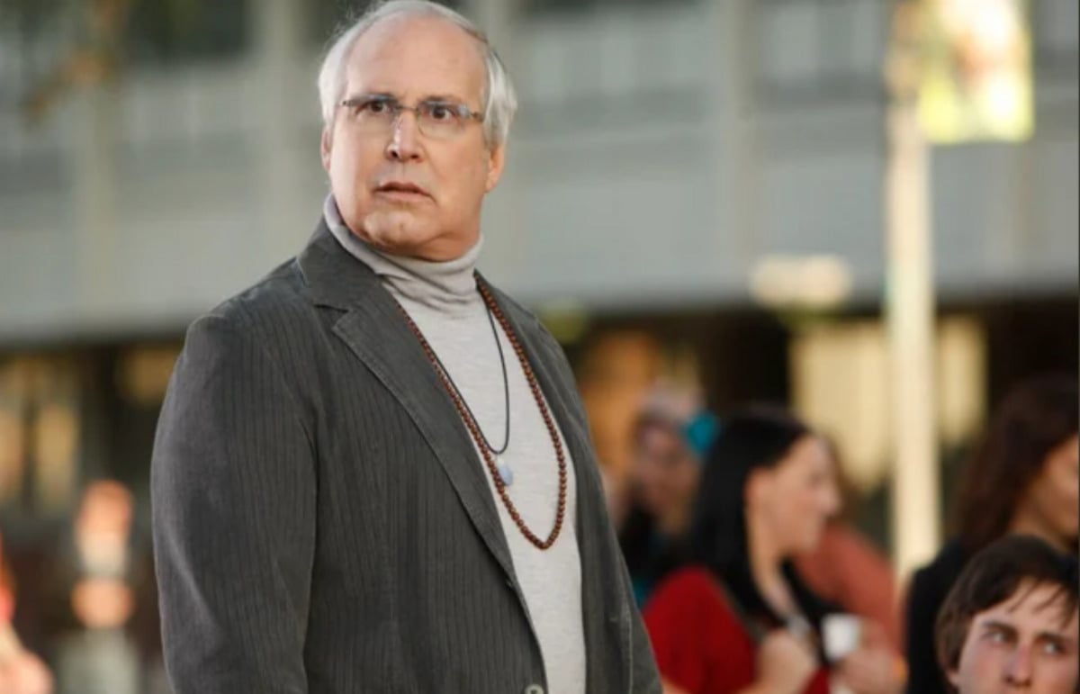 An older man with gray hair and glasses, wearing a gray suit jacket, turtleneck, and bead necklaces, stands outdoors with a surprised expression. People and a blurry building are in the background.