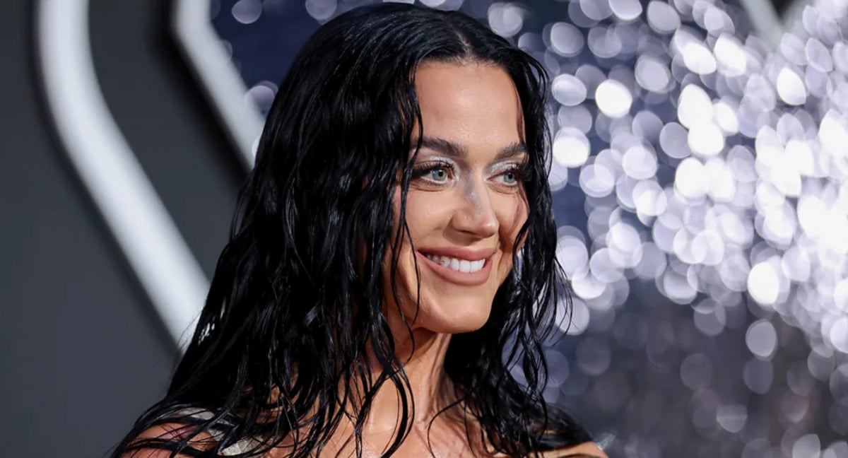 A woman with long, wet-look dark hair smiles while standing in front of a sparkling, out-of-focus background. She is wearing shimmery makeup and a strapless outfit.