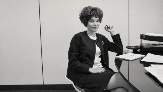 A woman with short hair in 1960s-style clothing sits at a desk, holding a cigarette. Papers and office supplies are on the desk. The photo is black and white. work, side hustle