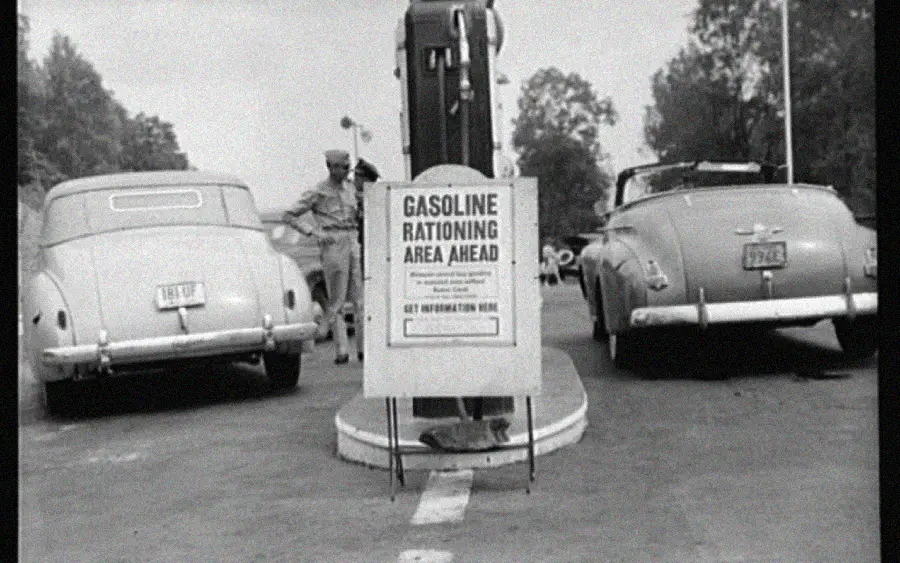 Two vintage cars parked at a gas station with a sign reading “Gasoline Rationing Area Ahead”; a uniformed attendant stands near the pump in the background. The scene appears to be from the 1940s.