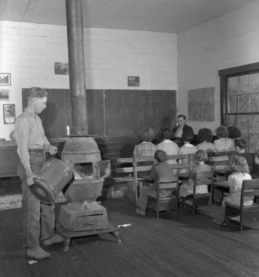 A black and white photo of a classroom where a boy tends a wood stove in the center, while a teacher sits at the front and students in rows face the blackboard, writing or listening. The room has wooden walls and large windows.