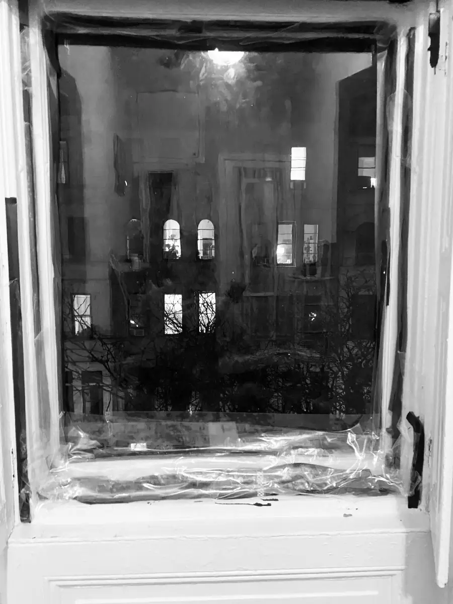 A black and white photo of a window covered in plastic, looking out onto a city building at night with illuminated windows and faint tree branches visible outside.