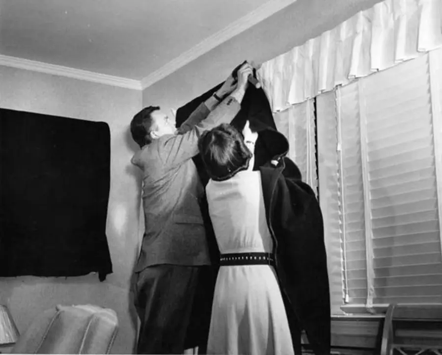 A man and woman hang a dark cloth over a window in a living room, partially covering the blinds. The room has light-colored walls, a curtain valance, and a cushioned chair in the corner.