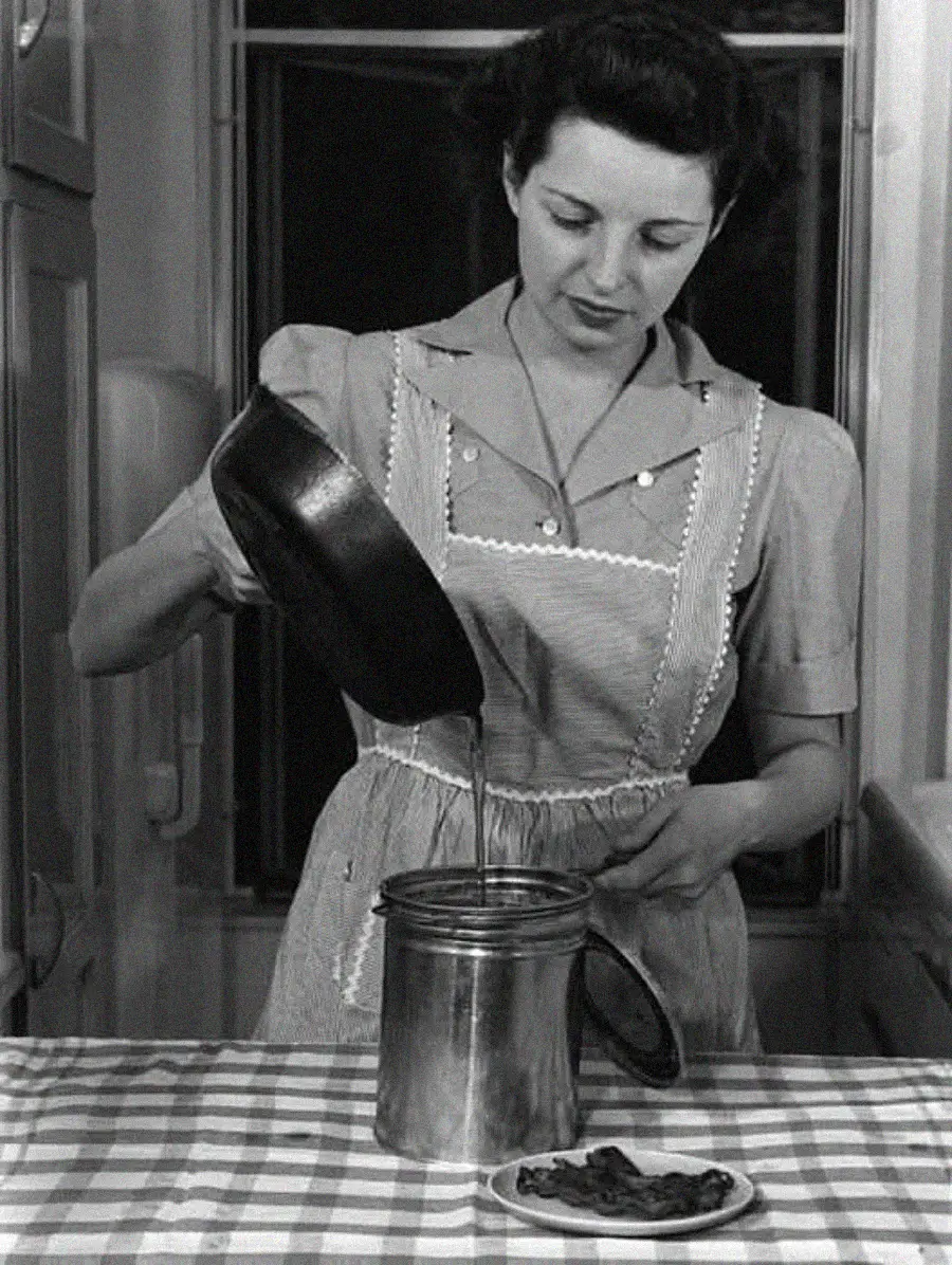 A woman in a vintage dress and apron pours liquid from a pan into a metal canister on a checkered tablecloth, with a small plate of food nearby.