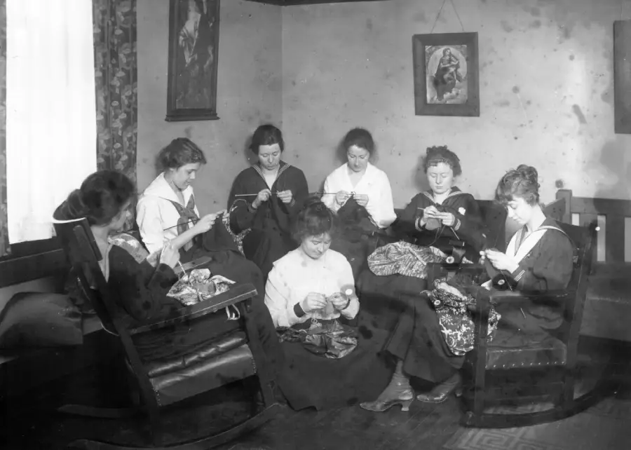 Seven women sit in a living room, focused on knitting. They are dressed in early 20th-century attire, and framed pictures hang on the walls behind them. Light enters through a curtained window to the left.