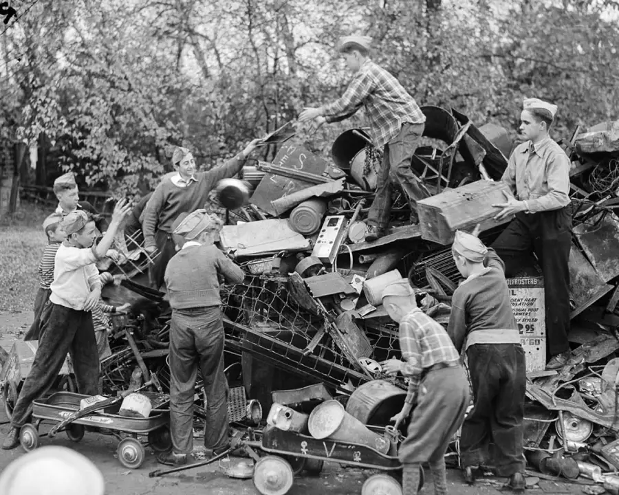 A group of boys, some in hats, collect and pile up scrap metal and old wagons outdoors, likely for a wartime scrap drive. Trees and foliage are visible in the background.