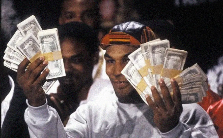 A man wearing a colorful cap and white shirt smiles and holds up several large bundles of cash in both hands, surrounded by people in the background.