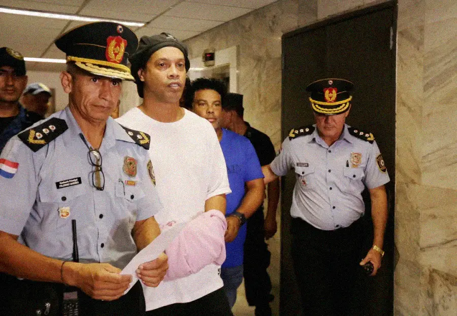 A man in a white shirt is escorted through a hallway by uniformed police officers. One officer leads the group holding papers, while others walk beside and behind the man. The setting appears to be indoors, possibly in a government building.