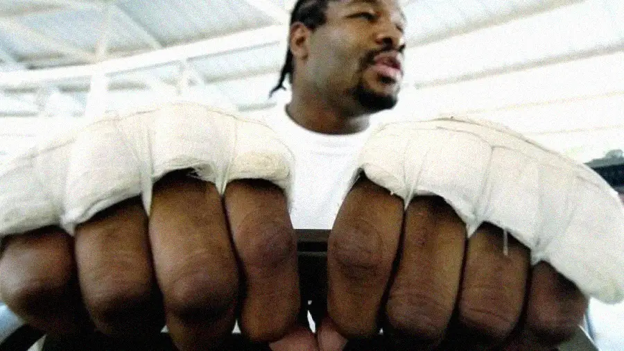 A close-up of a person’s fists wrapped in white athletic tape, held together and facing forward, with the person blurred in the background, possibly in a gym or training setting.