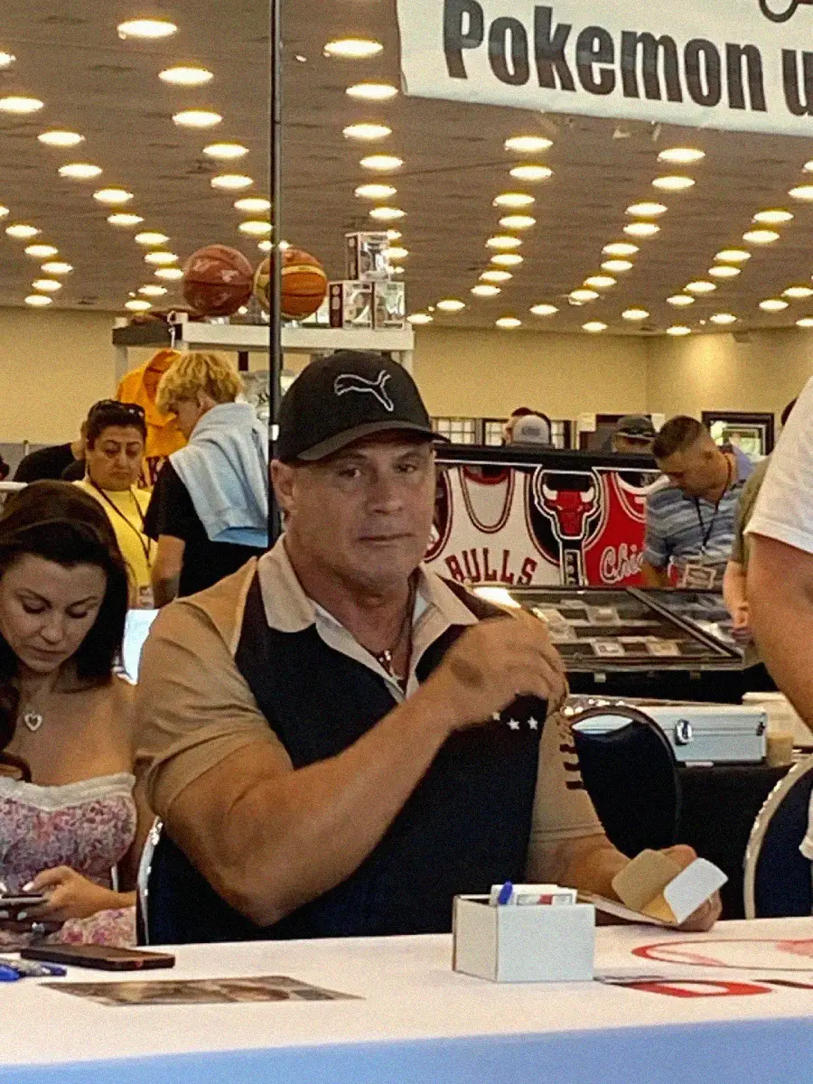 A man in a black Puma cap sits at a table signing items at a convention. People are around him, and sports memorabilia, including Chicago Bulls jerseys, is displayed in the background.
