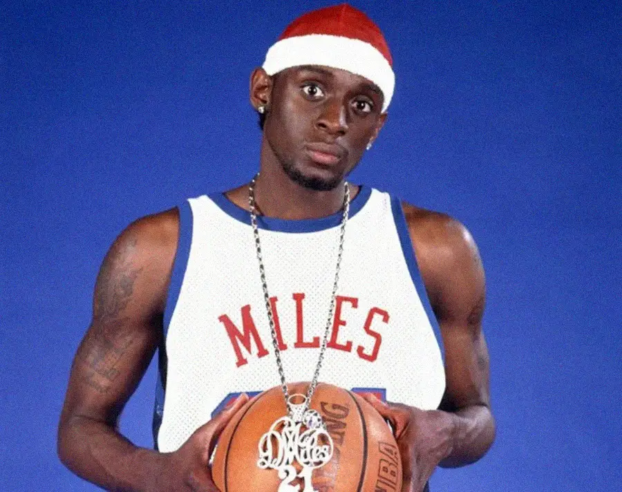 A basketball player wearing a white "Miles" jersey, red and white headband, and a large necklace, holding a basketball against a blue background.