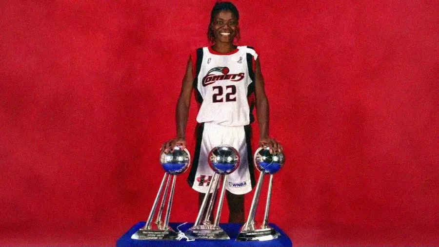 A basketball player in a Houston Comets uniform stands smiling with hands on three championship trophies, set against a red background.