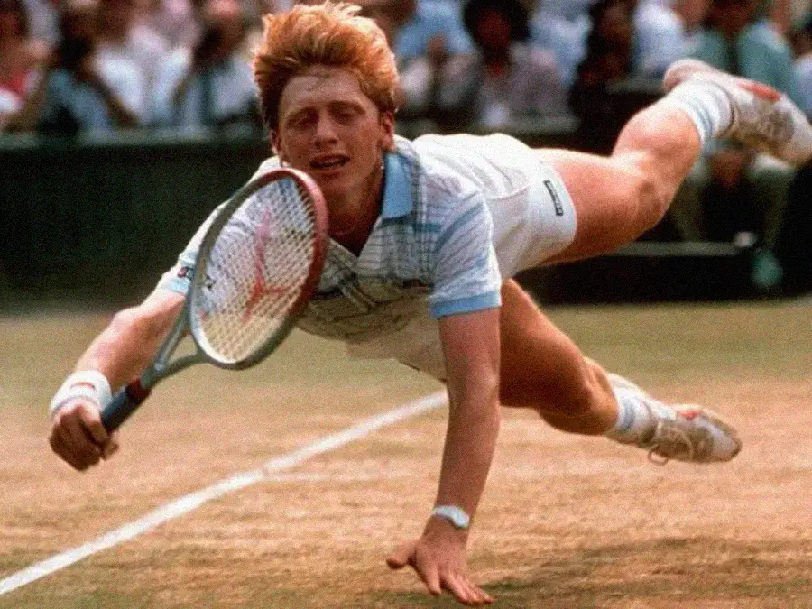 A tennis player in mid-air dives to reach a ball on a grass court, holding a racket with one hand and touching the ground with the other, showing intense effort and focus. Spectators watch in the background.