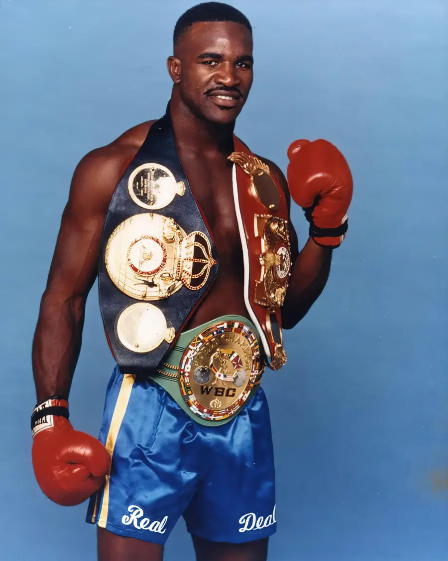A boxer wearing red gloves and blue shorts stands proudly, smiling, with multiple championship belts draped over his shoulders and waist against a plain blue background.