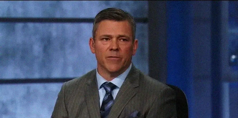 A man in a gray pinstripe suit and blue tie sits indoors against a blurred background with large window panels.