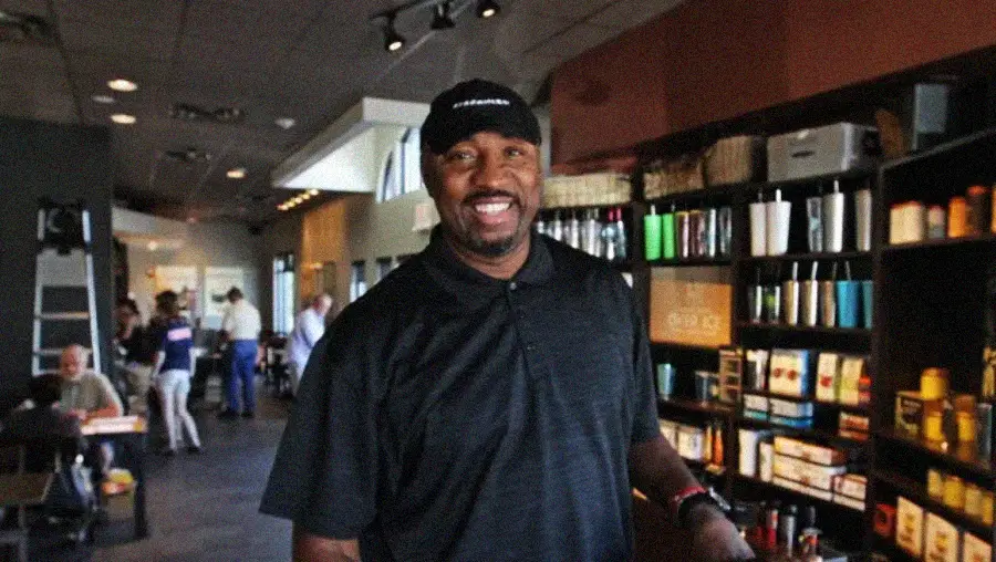 A man wearing a black polo shirt and cap smiles inside a coffee shop, with shelves of mugs and merchandise behind him and people sitting at tables in the background.