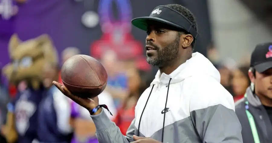 A man wearing a black visor and white Nike jacket holds a football in one hand, looking focused. There are blurred people and colorful backgrounds behind him, including a person in a mascot costume.