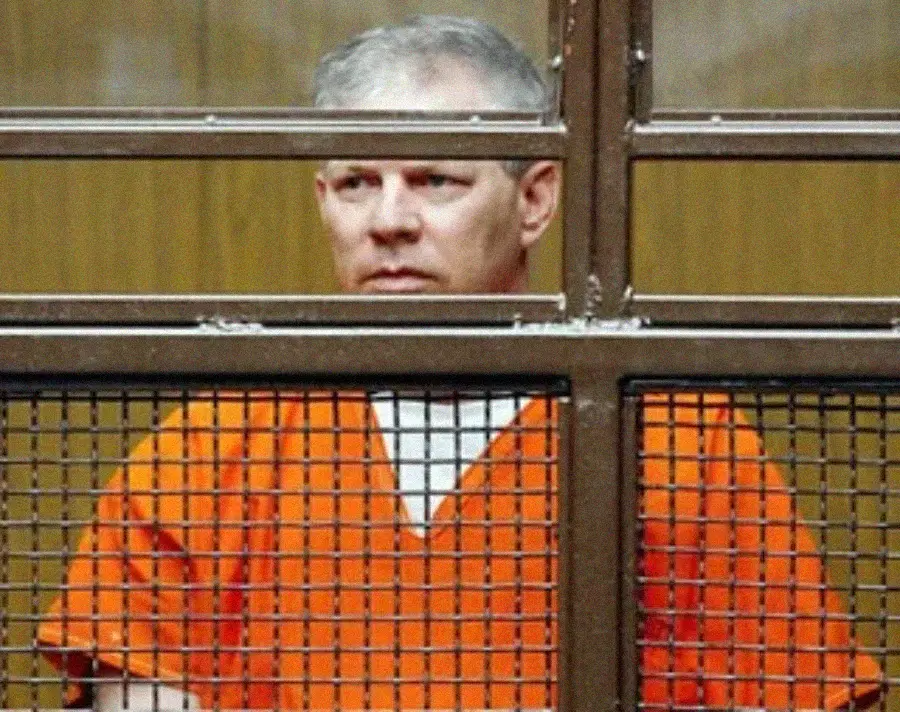 A man in an orange prison jumpsuit stands behind a metal cage and bars in a courtroom, looking forward with a neutral expression.